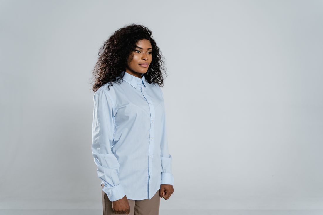 A woman in a light blue button-down shirt stands in three-quarter portrait against a plain neutral wall, calm and composed, facing slightly off-camera.