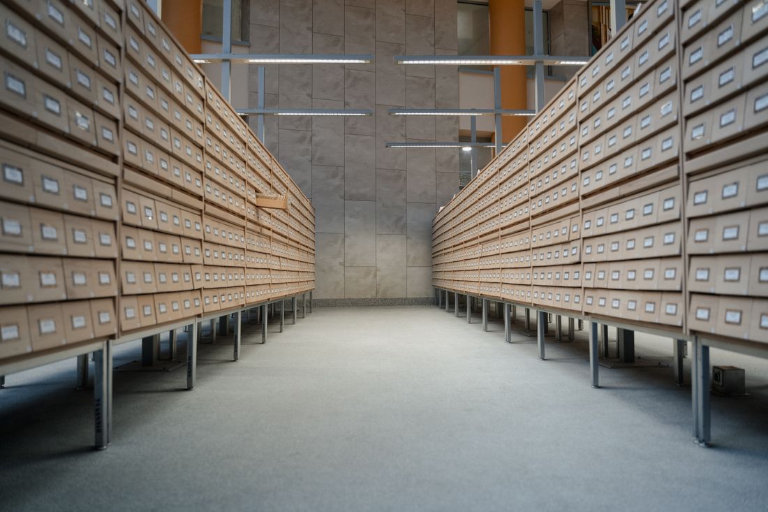 Rows of wooden archive storage drawers on shelving units receding down a bright, empty aisle, with no people present.