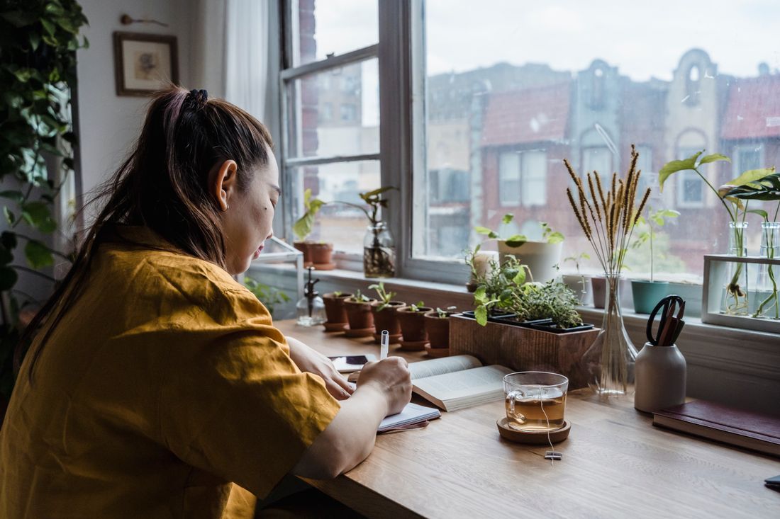 A person seated at a wooden desk by a bright window, writing in a notebook with a pen, surrounded by potted plants and an open book.