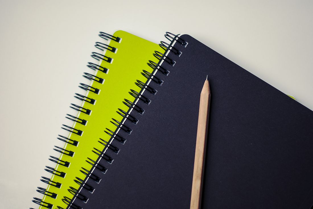 Two spiral-bound notebooks, one lime green and one navy, lying on a plain surface beside a sharpened wooden pencil.