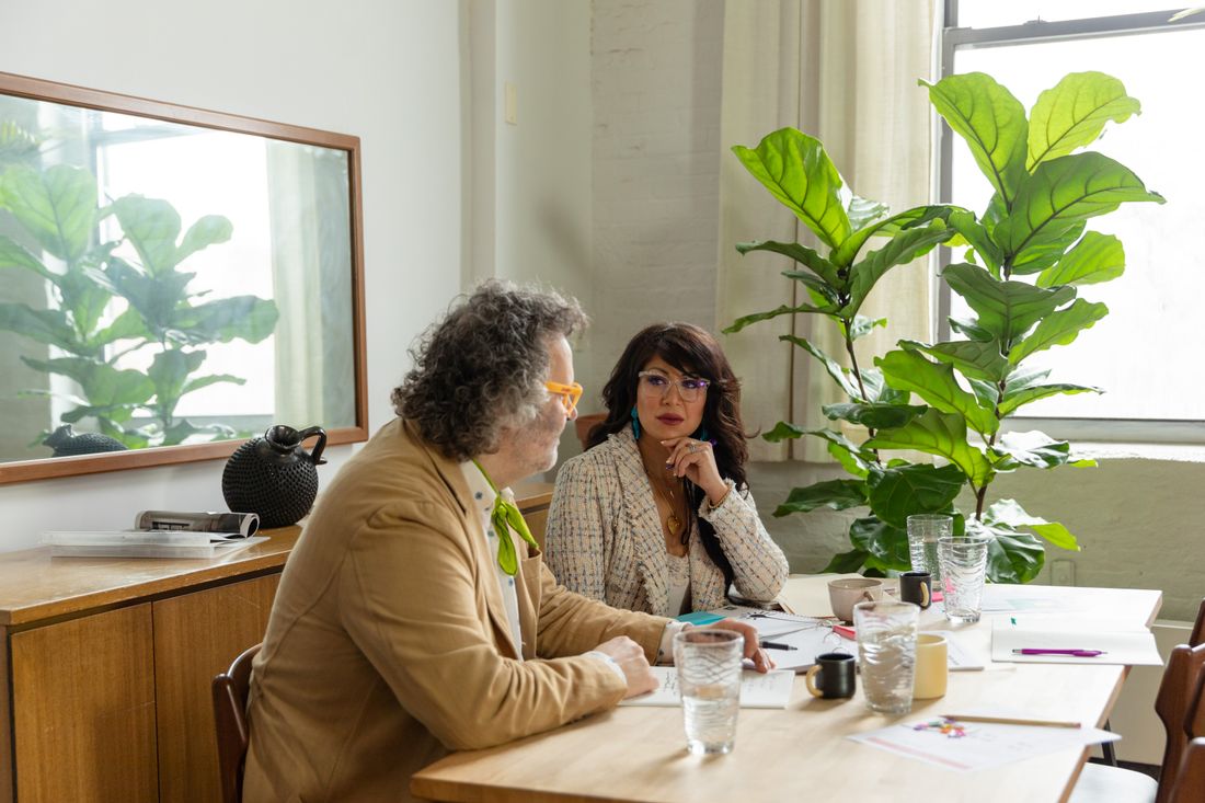 Two professionals sit across a small table in a bright, plant-filled office, talking with papers and drinks between them.