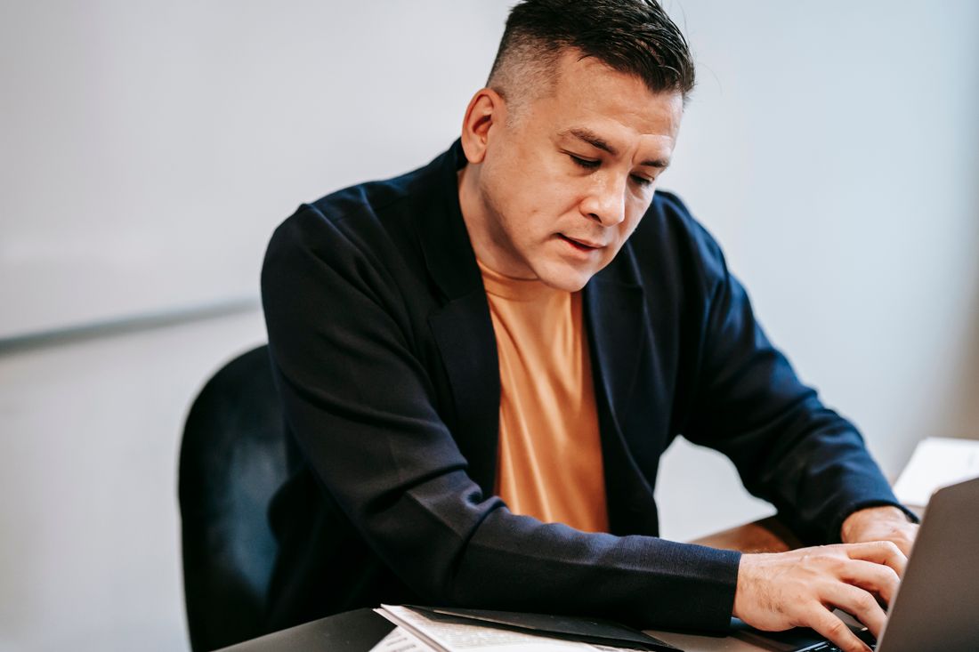 A man in a blazer works alone at a laptop against a plain bright wall.