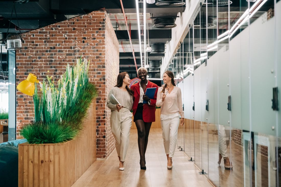 Three colleagues walk together through a modern office corridor with exposed brick and glass-panel walls, mid-conversation.
