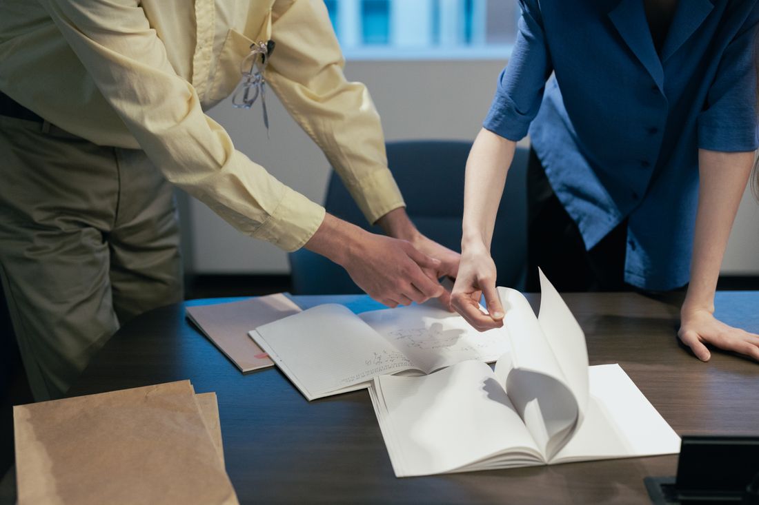 Two colleagues' hands examine an open notebook together at a wooden table during a focused conversation.