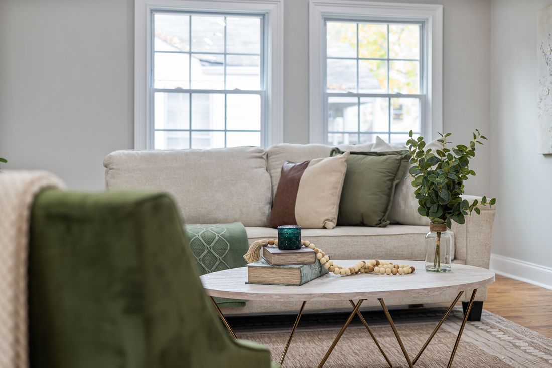 A furnished sitting area with a cream sofa, a green velvet armchair, textured cushions and a low round table, lit by daylight from two windows.