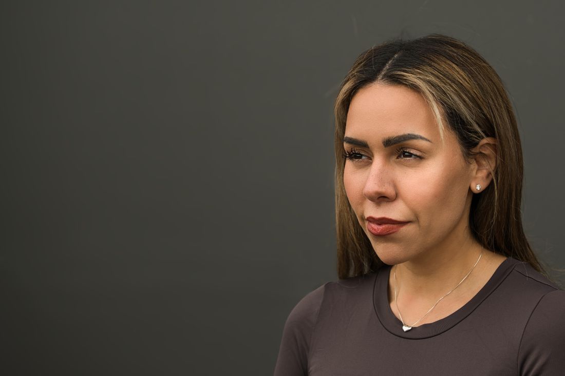 A head-and-shoulders portrait of a person in a soft taupe top against a plain dark background, turned slightly away from the camera.