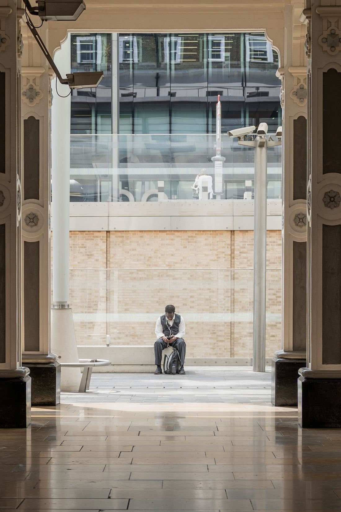 A man sits alone on a bench under a grand archway, checking his phone while he waits.