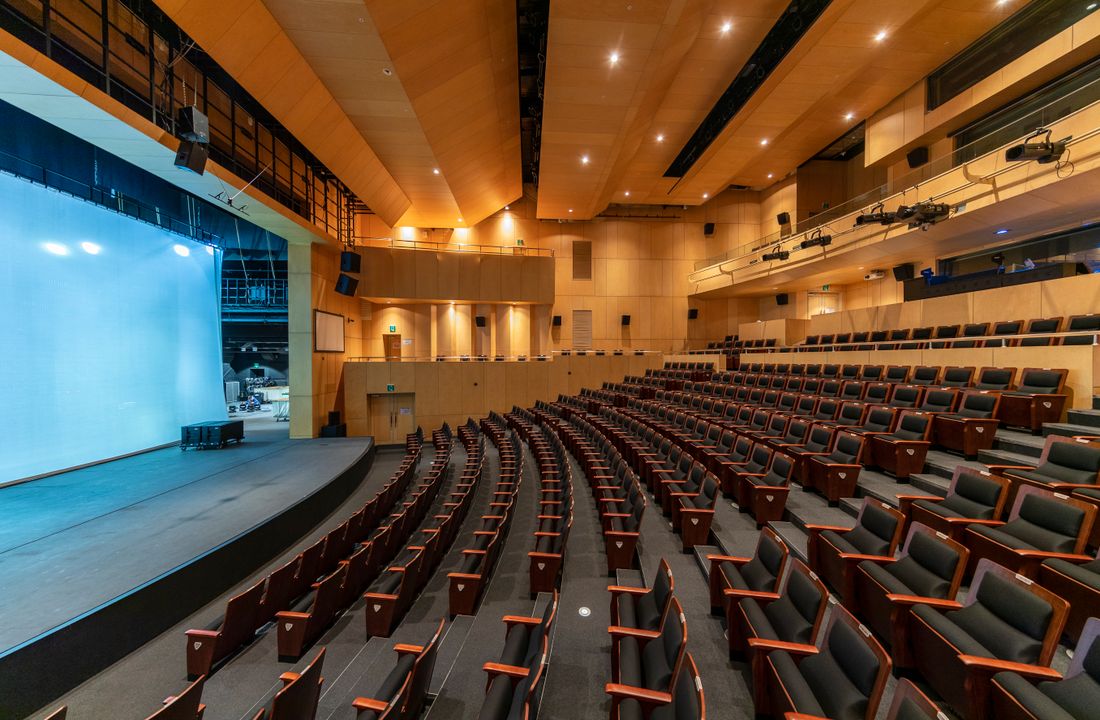 Rows of empty seats curve toward a low stage with a large blank blue-lit screen in a modern auditorium.