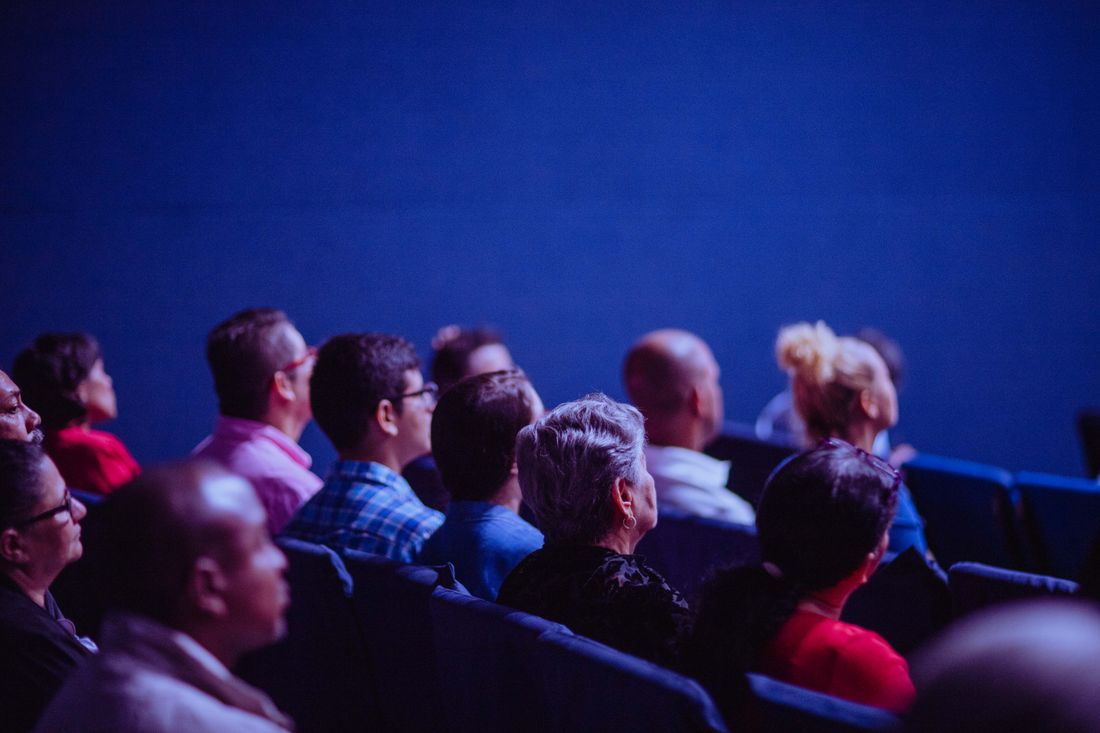 A seated audience viewed from behind, heads turned toward the front of a dim, blue-lit room.