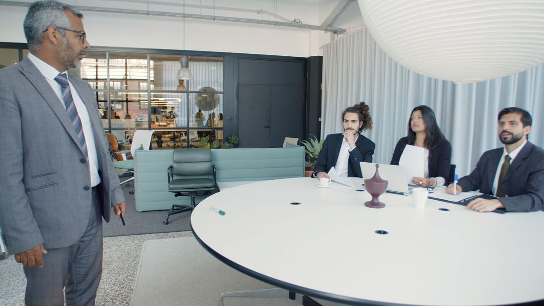 A standing executive addresses three seated colleagues in business attire around a round table in a modern meeting room.