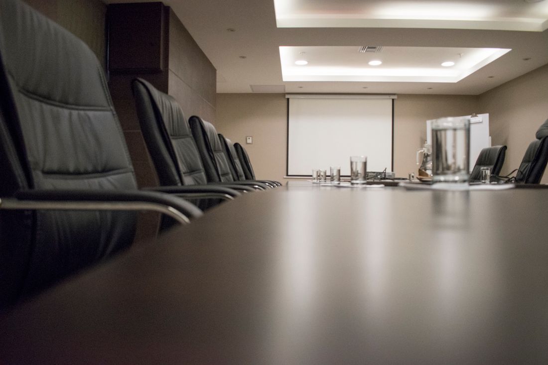An empty boardroom table lined with black leather chairs and water glasses set out, with a blank presentation screen in the background.