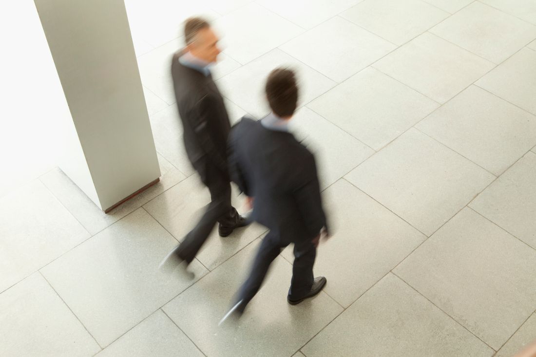 Two people in dark business suits walking briskly through a corridor, seen from above with motion blur across a tiled floor.