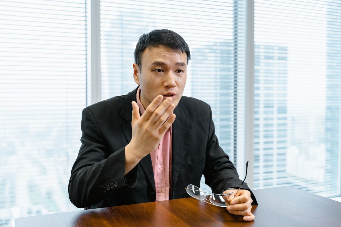 A professional sits at a wooden desk near a window, mid-conversation, gesturing while explaining a point during a one-on-one filmed interview.