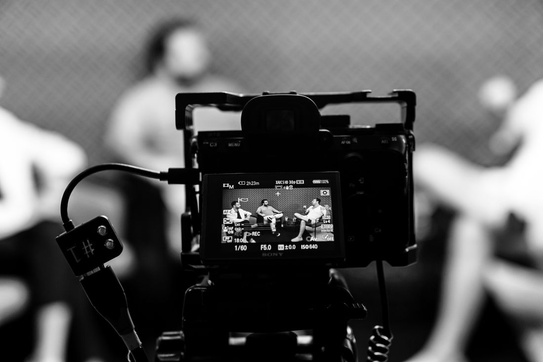 A camera's flip-out monitor shows a black-and-white view of an on-camera interview in progress, with the seated subjects visible out of focus in the background.