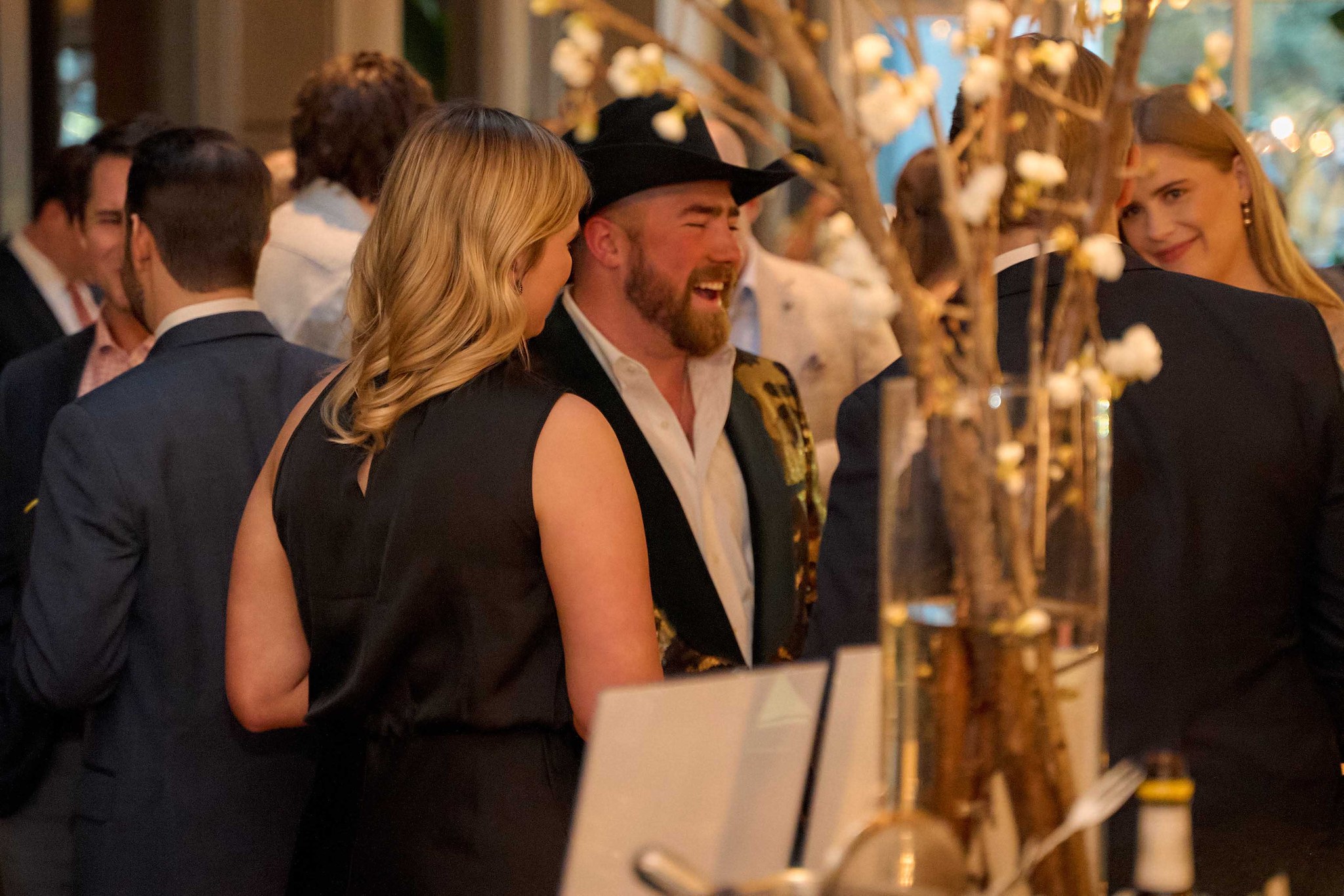 Man in black cowboy hat laughs while talking with woman at party with floral decor.