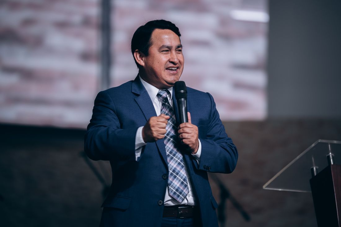 A speaker in a navy suit holds a microphone mid-sentence on stage, an acrylic lectern beside him and a lit screen behind.