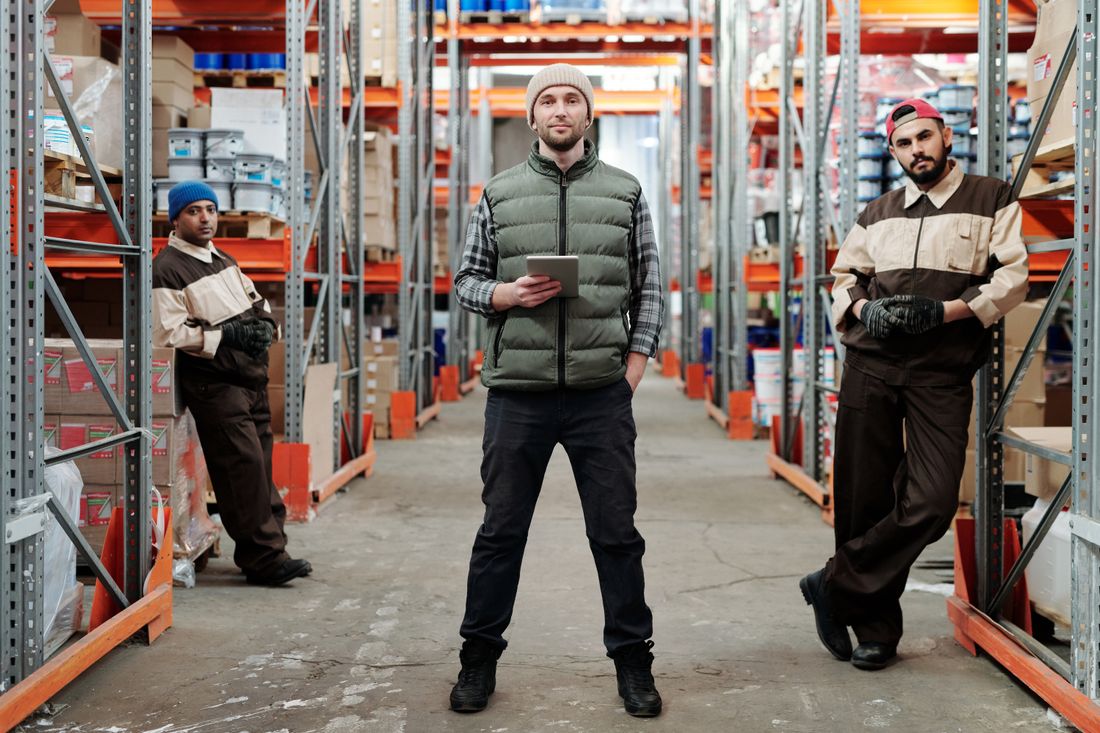 Three warehouse workers in work uniforms standing in a tall racking aisle, one holding a tablet.