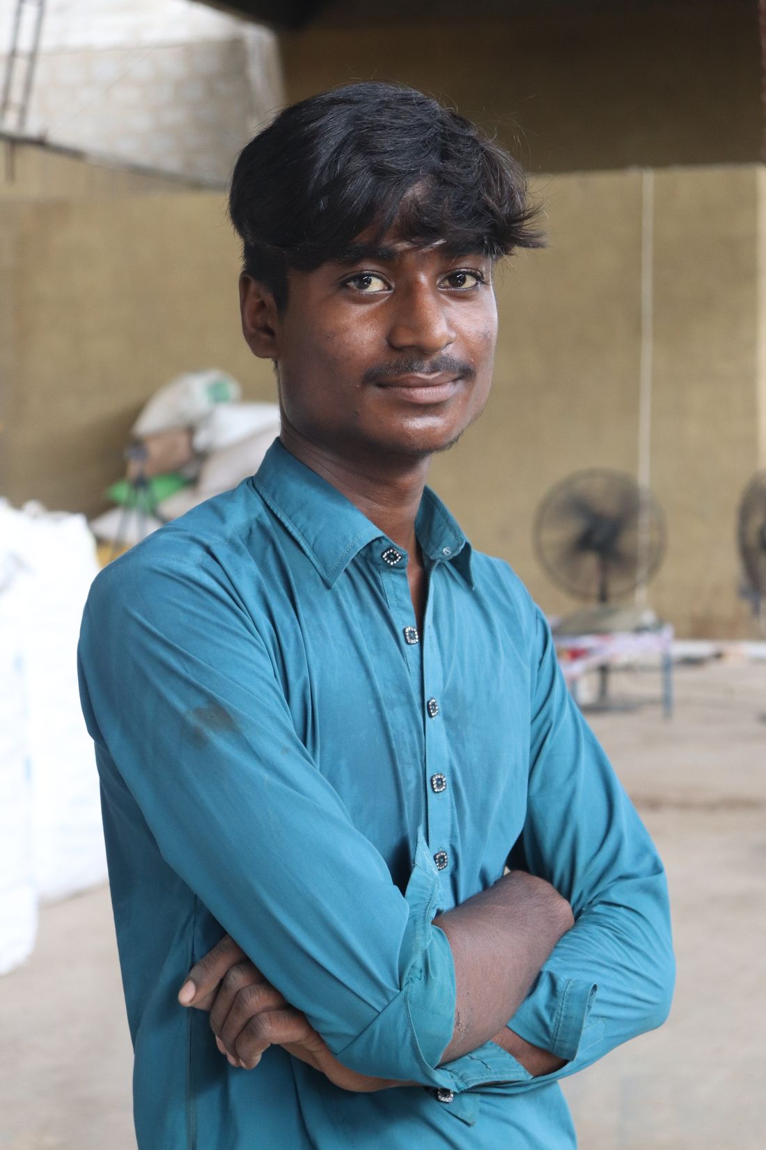 Portrait of a young worker in a collared work shirt, arms crossed, standing in front of a blurred industrial storage yard.