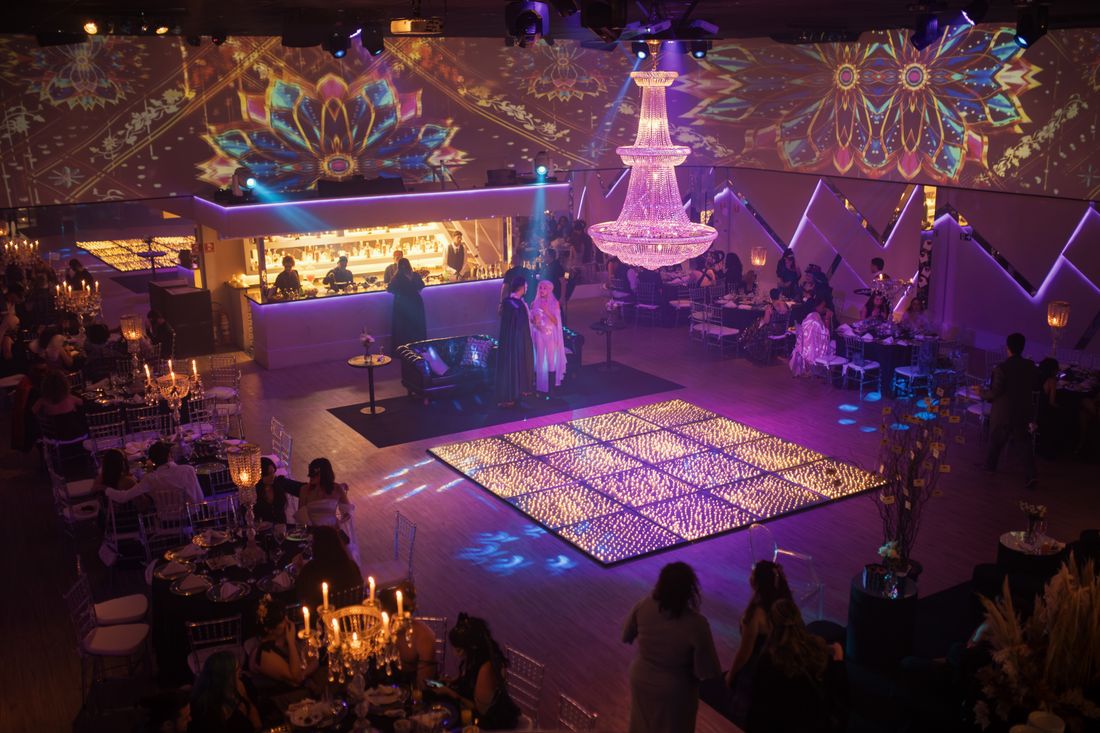 A dimly lit event space with colorful stage lighting, a glowing dance floor, and a crowd of guests under a large chandelier.