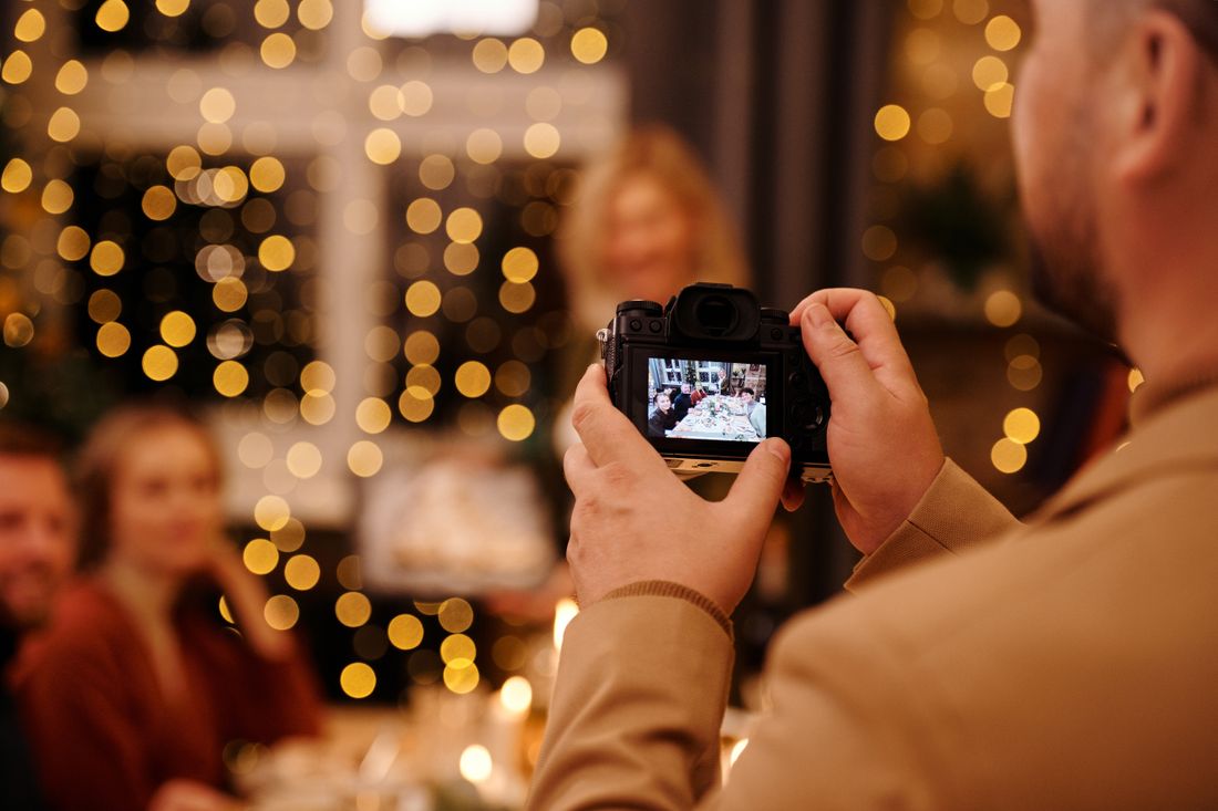 A guest photographs a warmly lit dinner gathering with a mirrorless camera, string lights glowing softly in the background.