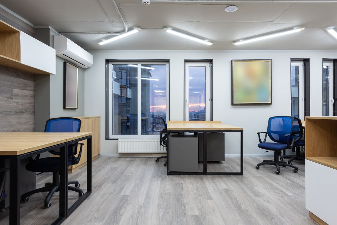 A modern office space with wood desks, blue chairs, and large windows looking out over the city at dusk.