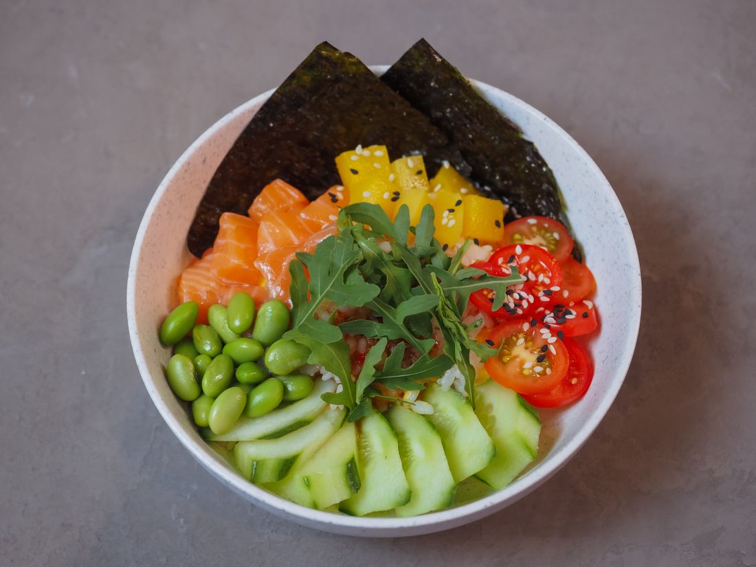 A poke-style bowl shot directly from overhead, filled with salmon, cucumber ribbons, edamame, mango, cherry tomato, and nori, centered against a plain neutral surface.