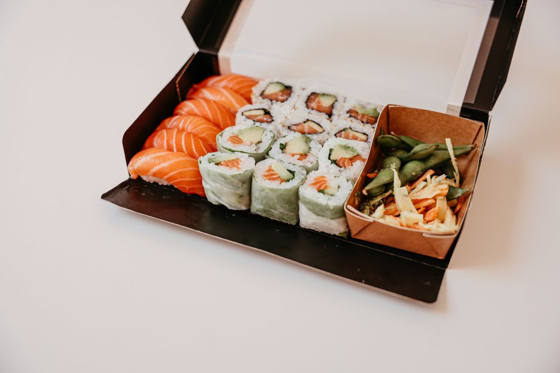 An open black clamshell takeout container on a plain white surface, shot at a three-quarter angle, holding sushi rolls, nigiri, and a small side of edamame slaw.
