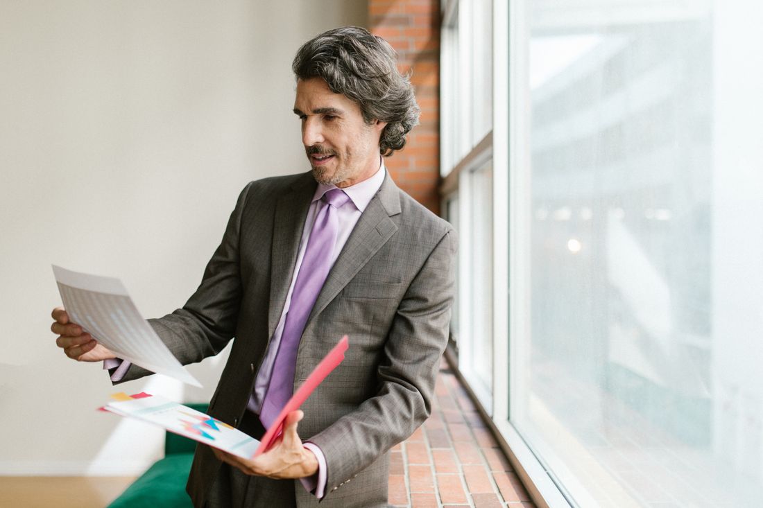 A professional in a gray suit stands near a large office window in natural daylight, reviewing a colorful printed chart and a document held in his other hand.