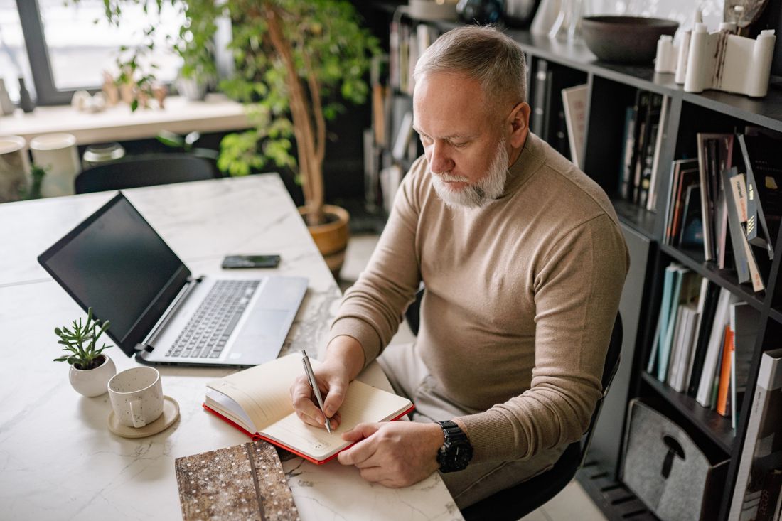 A bearded professional in a sweater sits at a marble desk writing in a notebook, with a closed laptop, a coffee cup, and a bookshelf in the background.