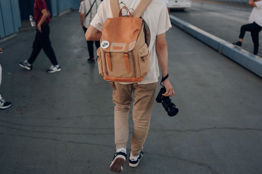 A person walking away down a city street carrying a camera in one hand and a travel backpack over their shoulders, seen from behind.
