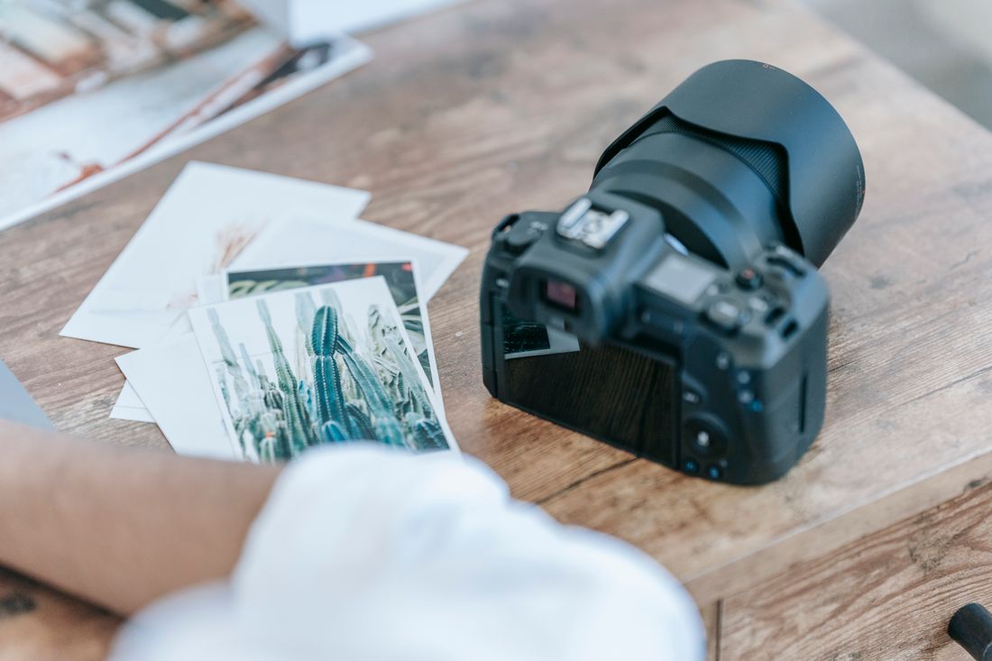 A camera resting on a wooden table next to a small stack of printed photographs, with a person's hand visible at the edge of the frame.