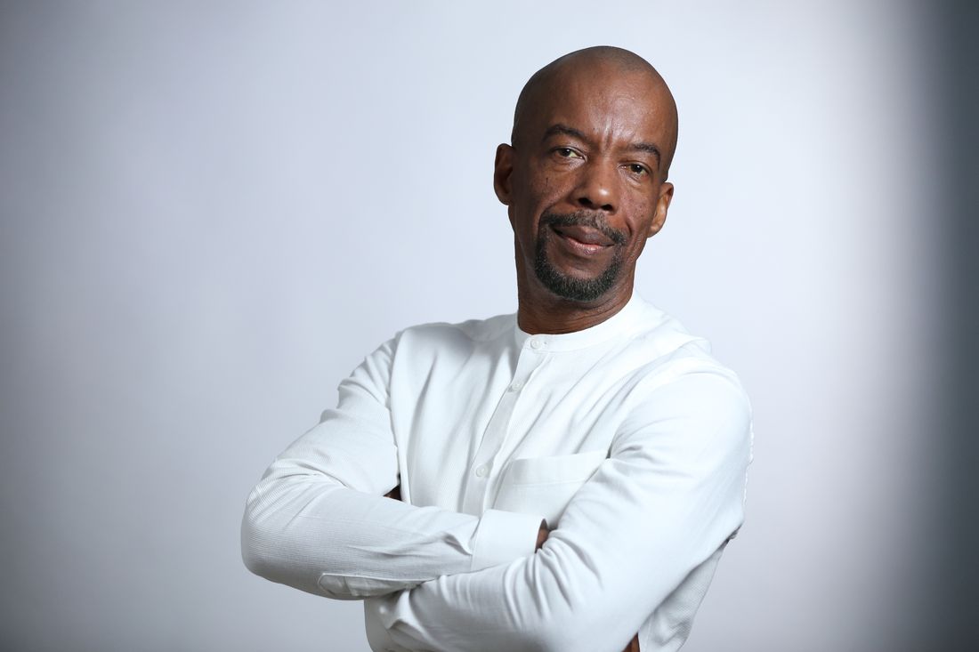 A bald man in a white collarless shirt stands with arms crossed against a plain gray studio backdrop, looking at the camera.