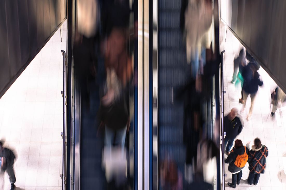 An overhead view of a busy indoor escalator area with several people in motion, carrying bags and backpacks, blurred by movement.