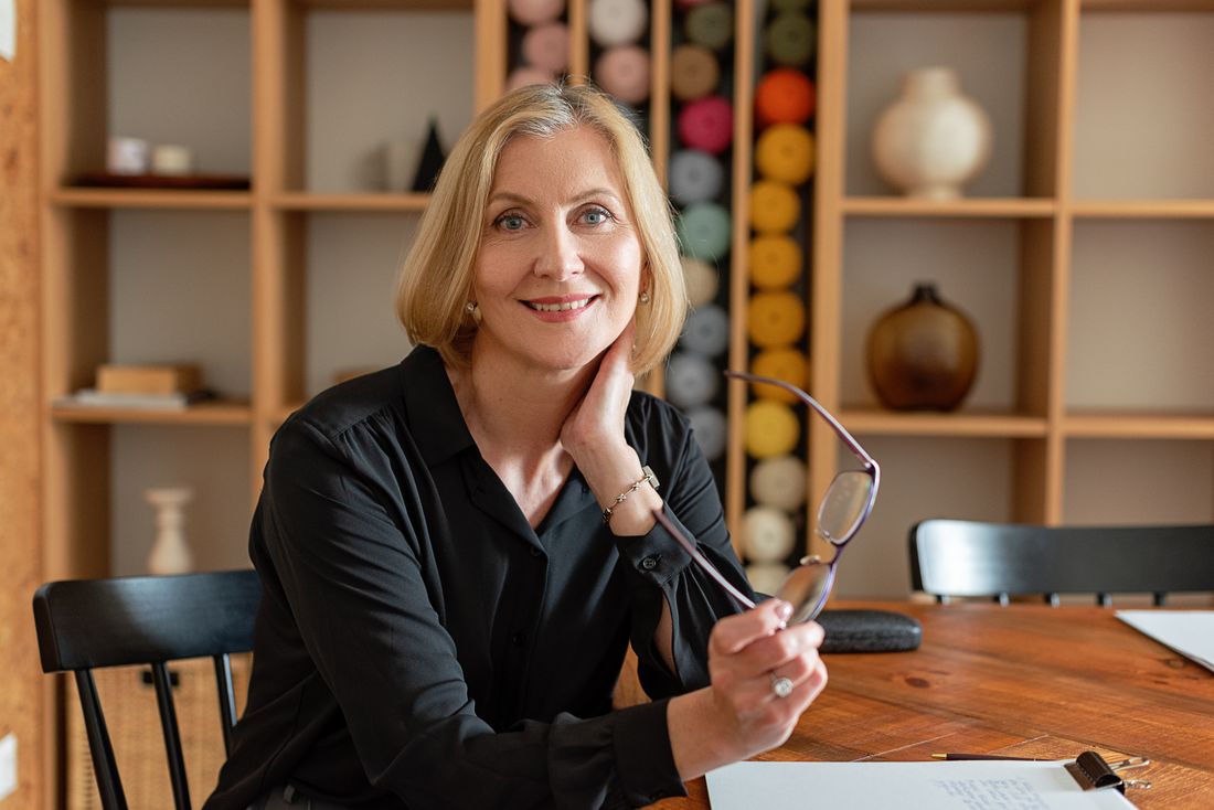 A woman in a black blouse sits at a wooden table holding her glasses, smiling warmly, with shelves of pottery and thread spools behind her.