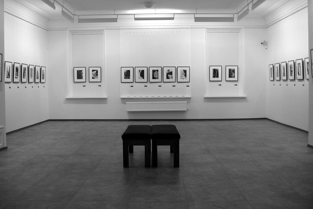 A quiet gallery room with plain white walls, rows of small framed black-and-white photographs, and two low stools on the floor.