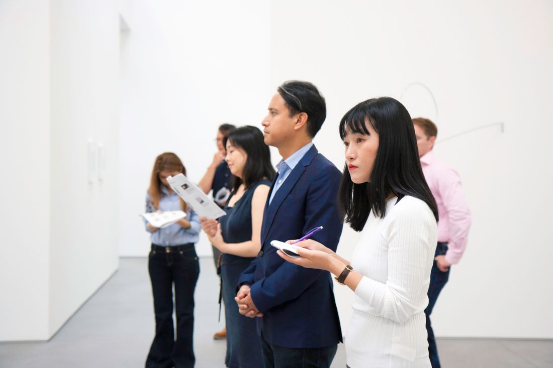 A small group of well-dressed visitors at a bright, minimalist gallery opening, some holding exhibition guides as they look around the room.