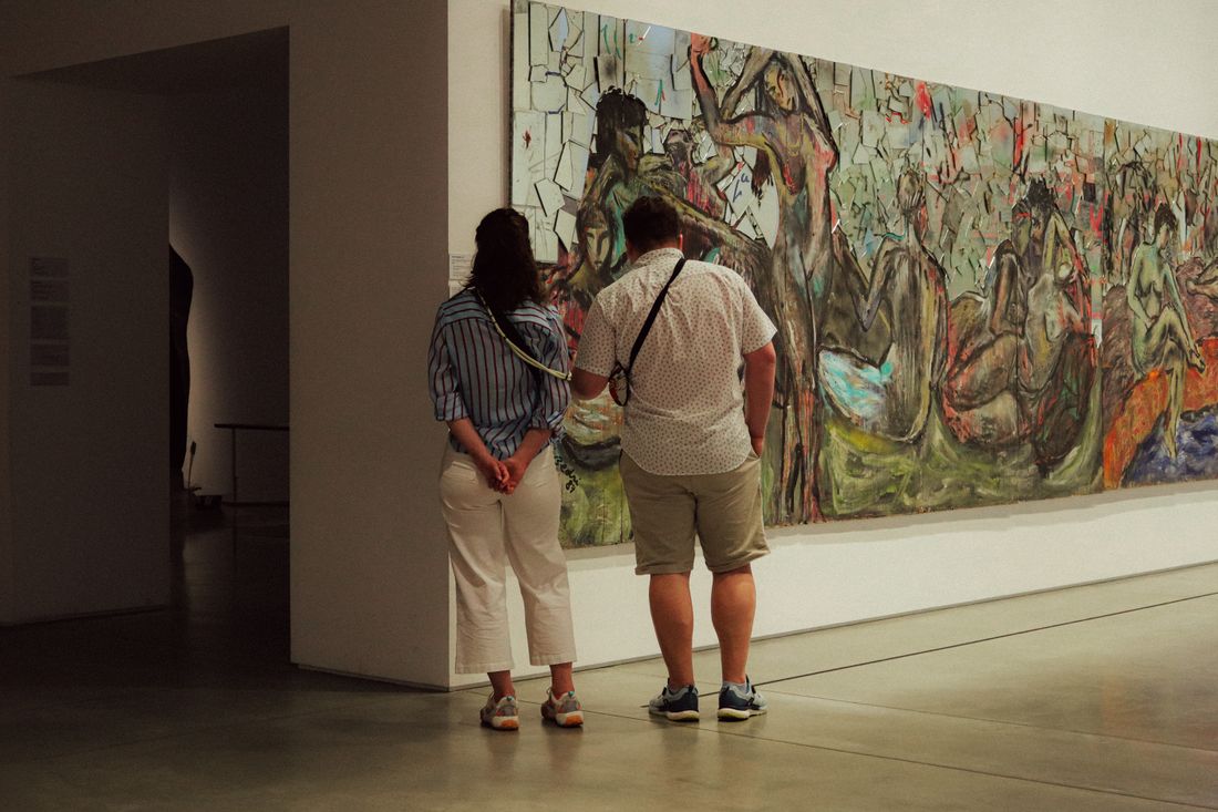 Two visitors seen from behind, standing close together as they view a large abstract painting on a gallery wall.