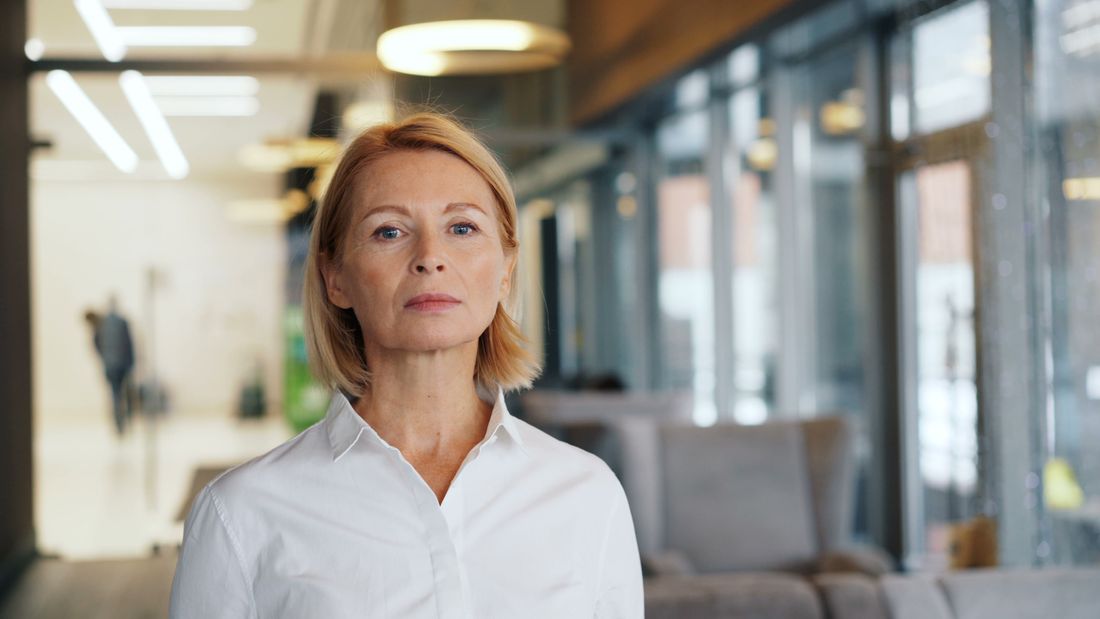 A confident, well-dressed woman stands for a portrait in a bright office hallway, natural light from windows behind her.