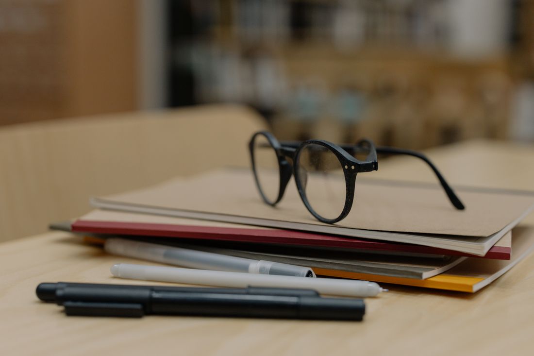 Closed notebooks and folders stacked on a light wood table beside a pair of reading glasses and several pens.