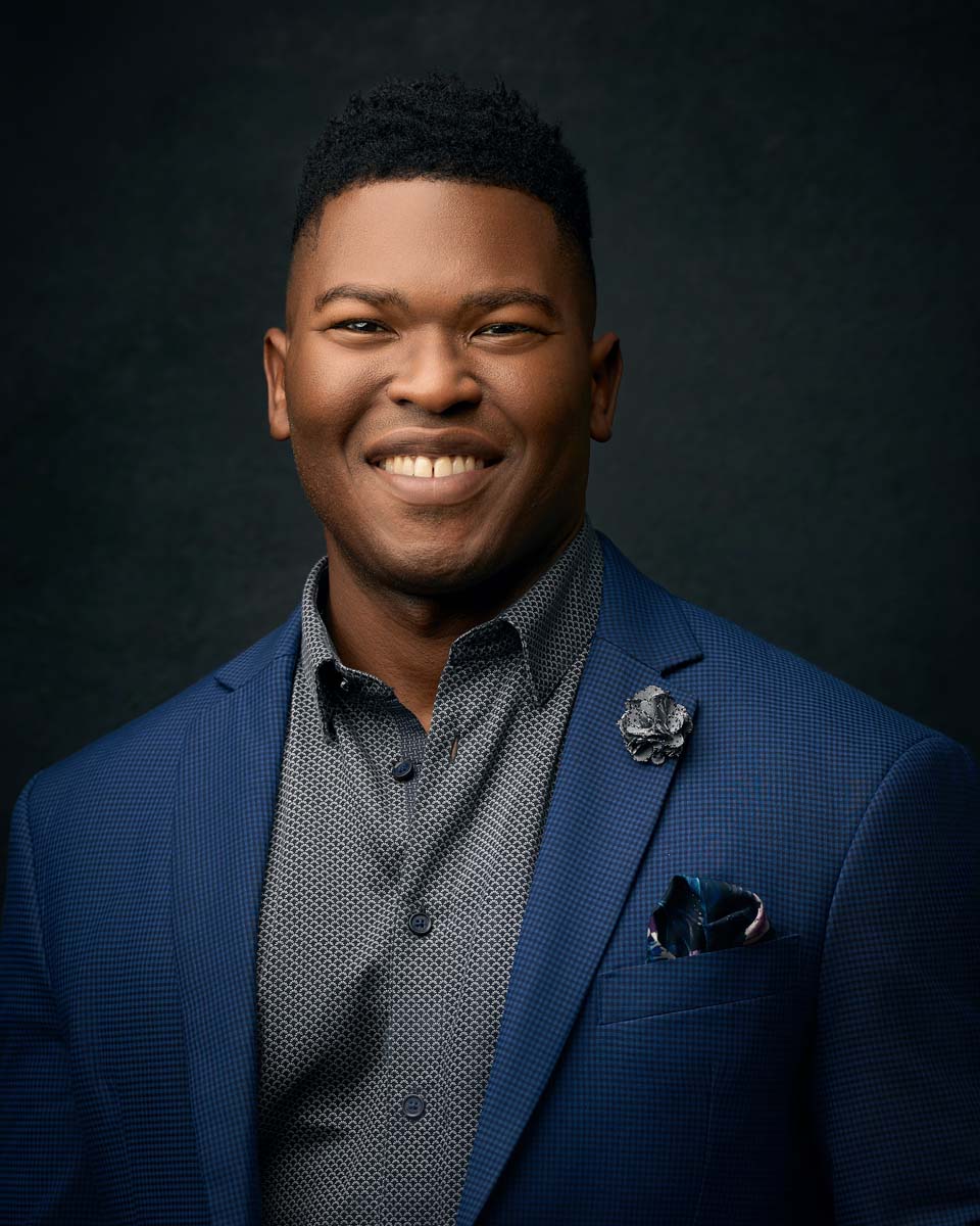 Smiling man in blue suit with pocket square poses for studio headshot against dark background.