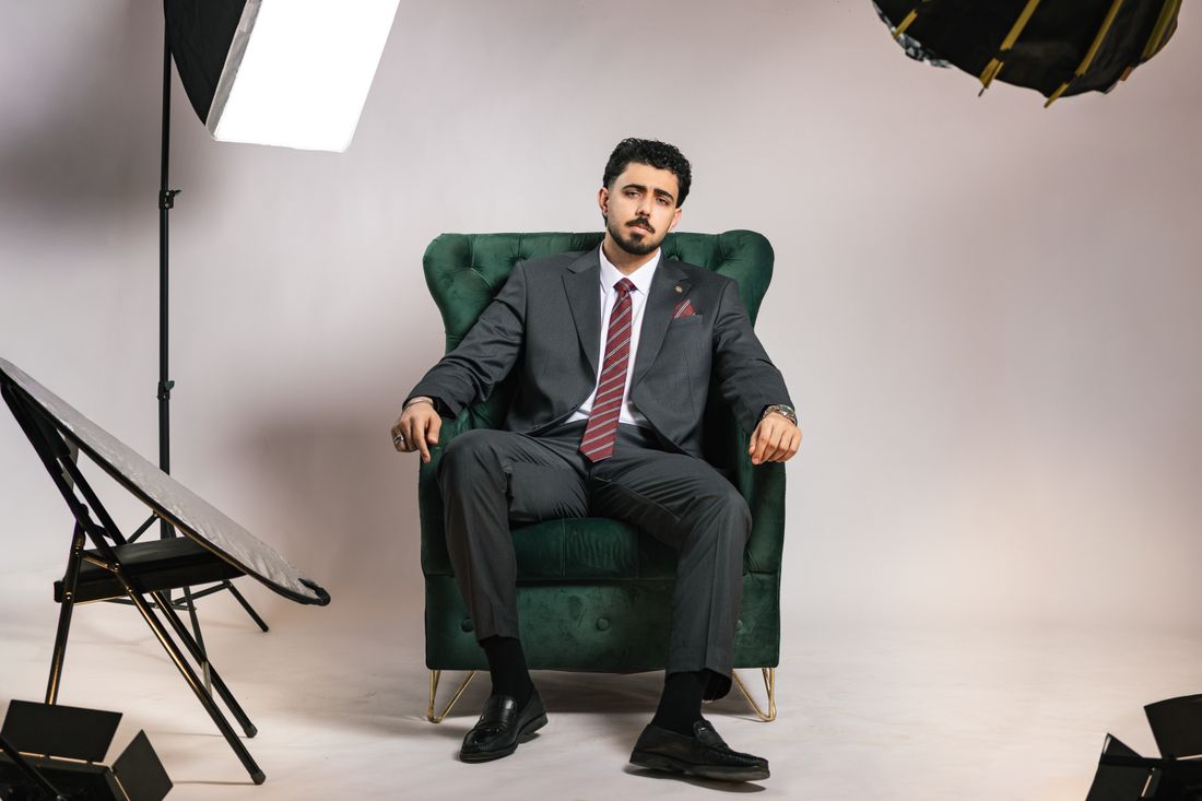A businessman in a suit sitting for a formal portrait session in a photography studio, lighting equipment visible on either side.