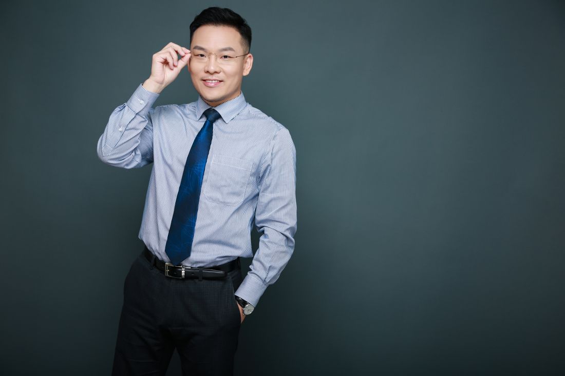 A man in a light blue dress shirt and dark tie stands against a plain dark gray wall, adjusting his glasses with a calm smile.