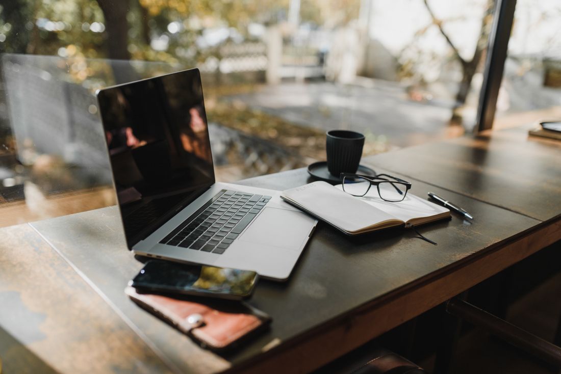 A laptop, notebook and glasses on a wooden table beside a window in a quiet workspace