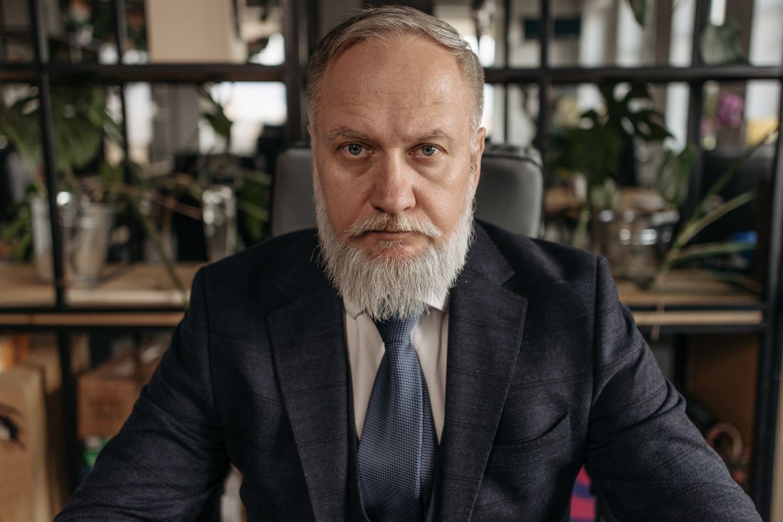 A mature, gray-bearded man in a dark suit and tie, arms uncrossed, looking directly at the camera with a composed, serious expression against a softly blurred backdrop.