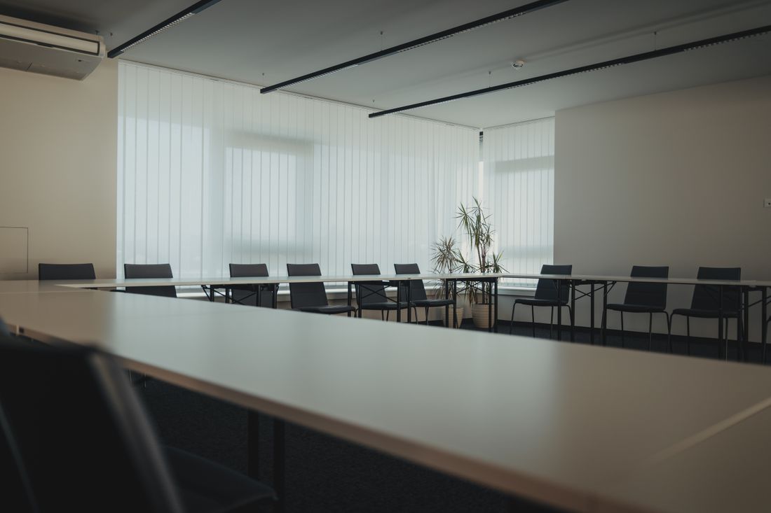 An empty meeting room with tables arranged in a U-shape, black chairs, sheer curtains diffusing daylight, and a potted plant in the corner.