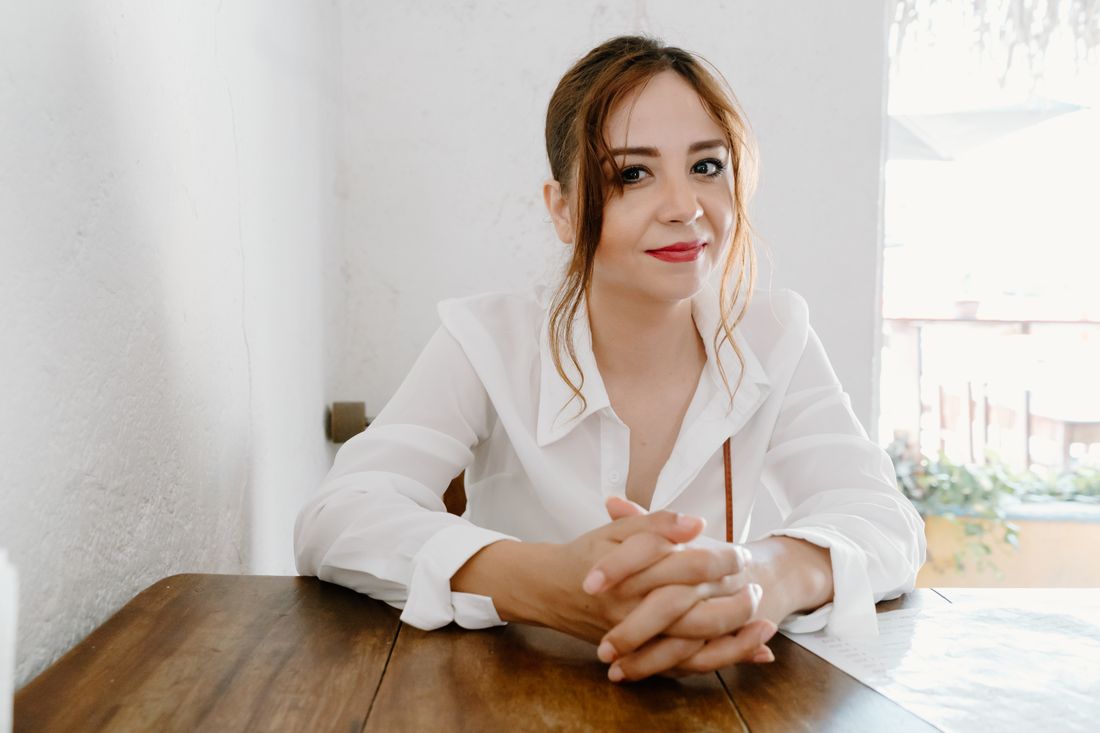 A woman in a white blouse sits at a wooden table in a plain, sunlit room, hands clasped, smiling gently at the camera.