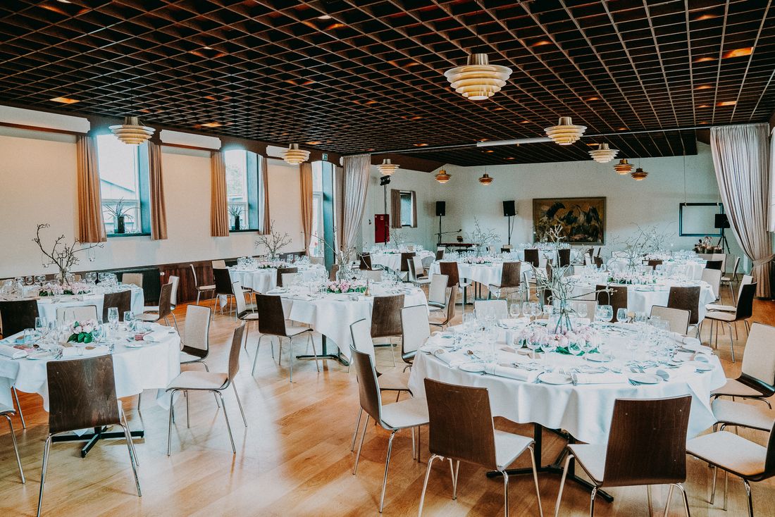 A bright hall set with round tables, white linens, and wooden chairs, with morning daylight coming through tall windows.