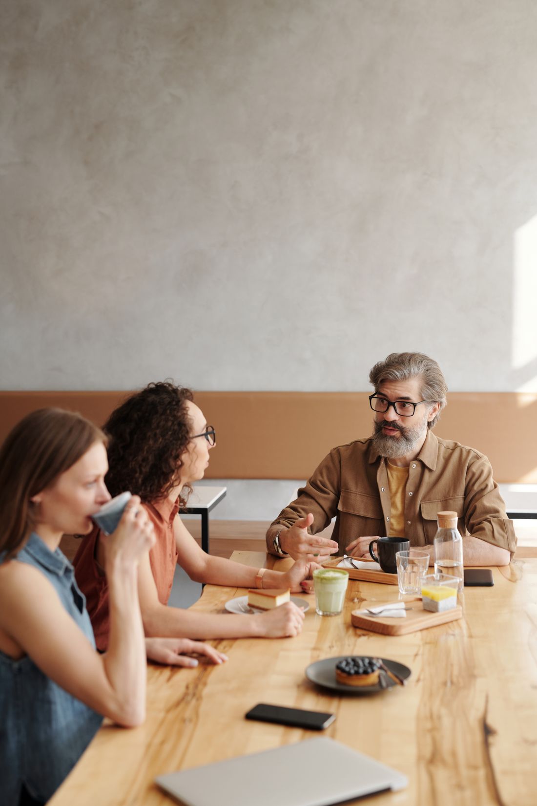 A small group of adults in casual-professional clothing sit together at a wooden table, talking warmly over coffee and pastries in daylight.