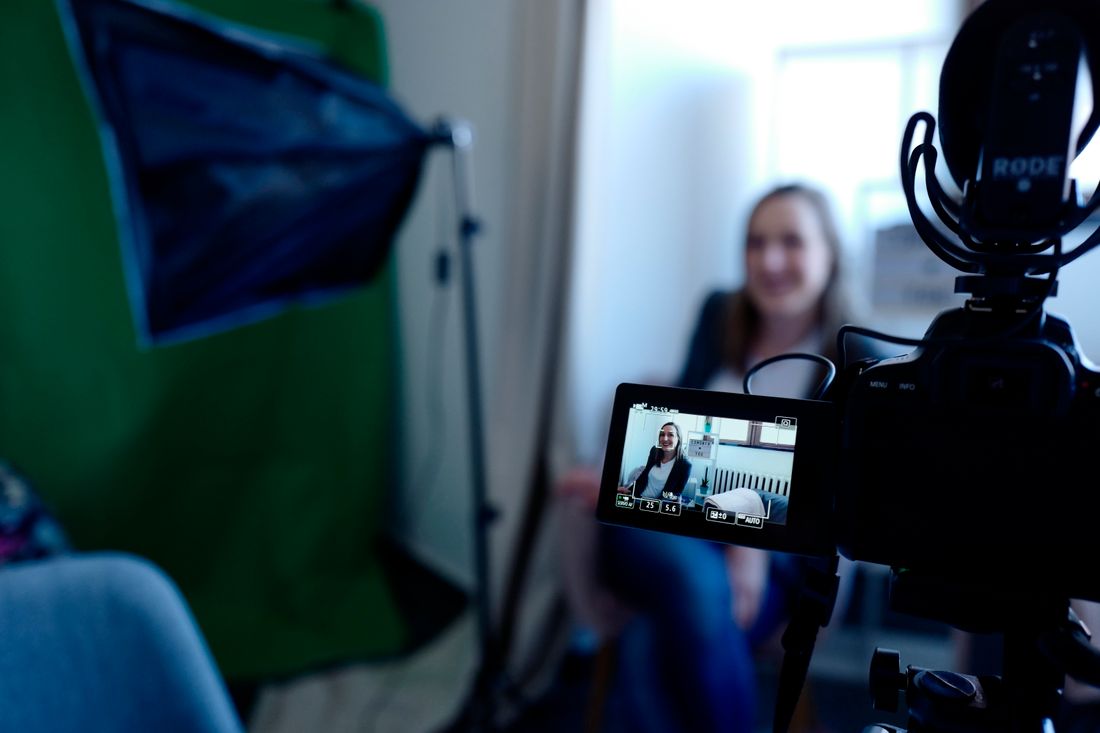 A video camera on a tripod records an interview subject seated in front of a softbox light, monitor showing the framed shot.