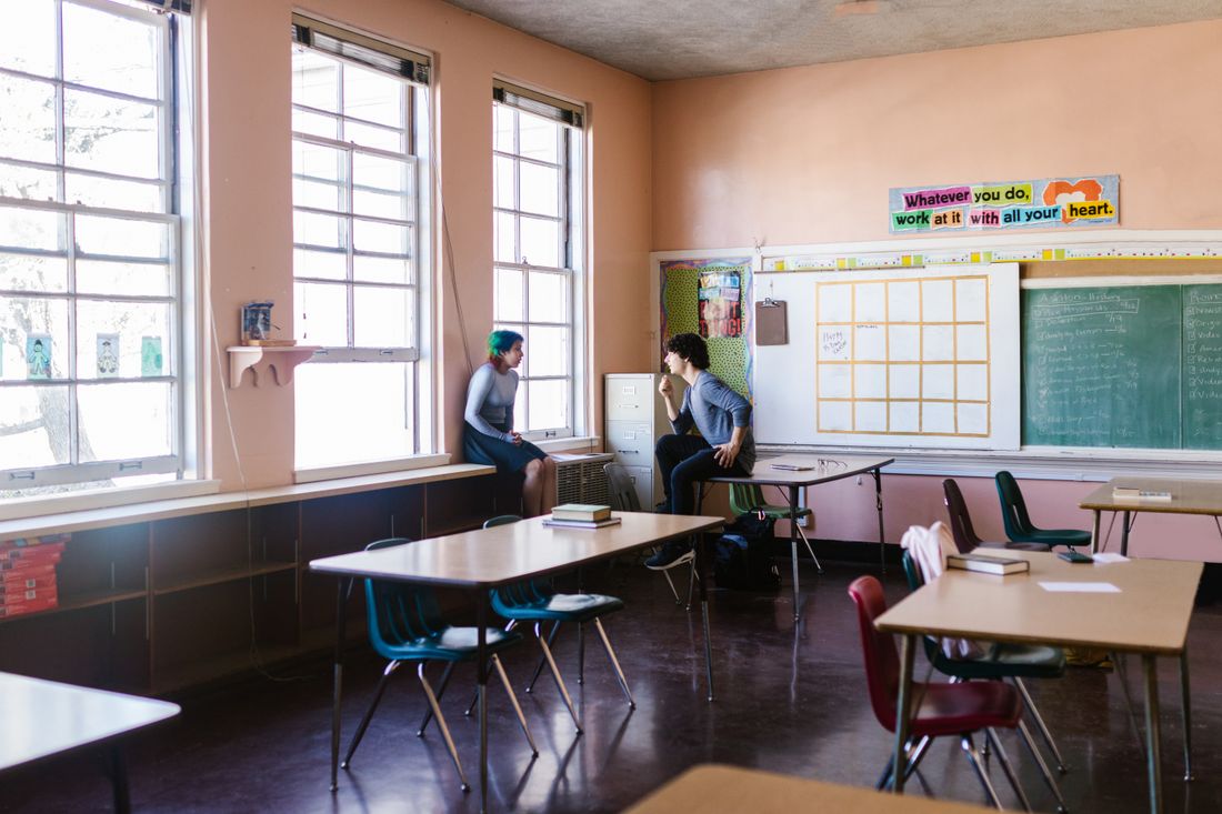 Two people talk quietly near a window in an empty classroom-style program space with desks and a chalkboard.